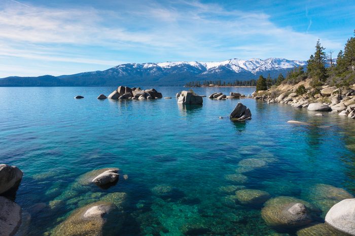 Clear turquoise water with submerged rocks reflects sunlit sky, framed by rocky shoreline and snow-capped mountains in the distance.