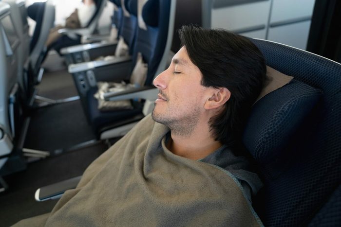 A person sleeps under a blanket, reclining in an airplane seat, with other passengers and seats visible in the background.