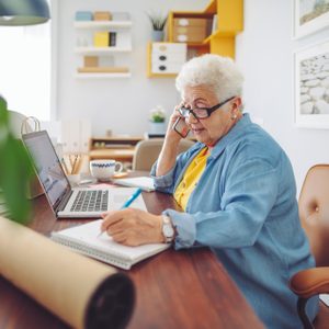 Elderly woman talks on phone, writes notes in a notebook at a desk with a laptop, in a home office setting.