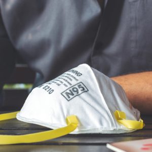 N95 mask resting on a table, labeled with text, beside a person wearing a dark shirt, in an indoor setting.
