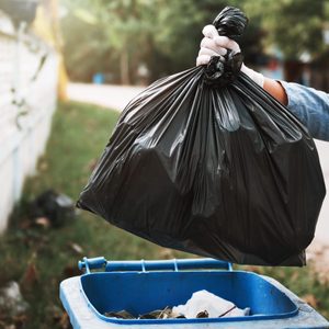 Gloved hand lifts black garbage bag over blue trash can, outdoors near a white fence and green foliage.
