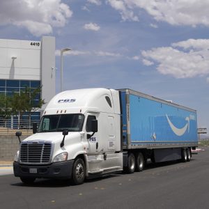 A semi-truck with a blue Amazon Prime trailer drives past a modern building numbered 4410 under a partly cloudy sky.