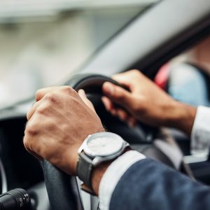 Hands grip a steering wheel inside a car, with a wristwatch visible, suggesting a focus on driving in a vehicle's interior environment.