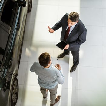 Two men converse near a parked car; one holds a tablet, using gestures in a bright, tiled showroom.