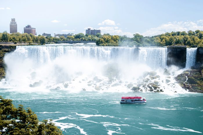 A boat tours near large waterfalls amid mist, with a backdrop of buildings and green trees under a bright sky.
