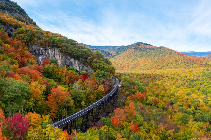 Bridge spans colorful autumn forest, surrounded by rolling mountains under a bright blue sky.