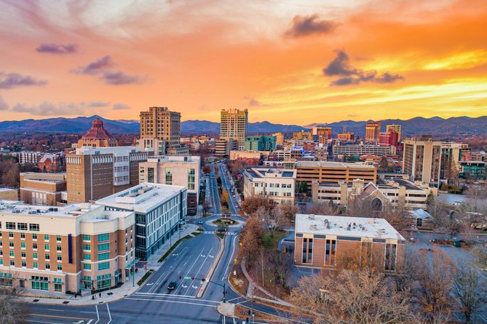 Buildings stand quietly under a vibrant orange sunset, with a cityscape foreground and distant mountains framing the horizon.