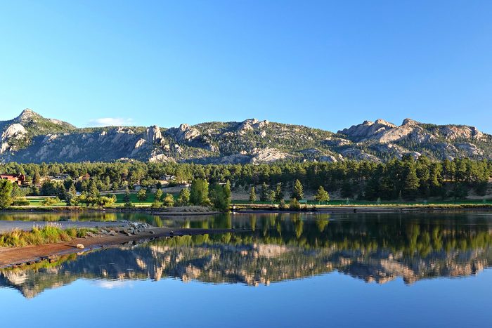 Mountains reflect on a calm lake, surrounded by a forest and clear blue sky.