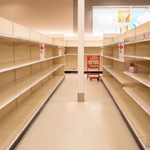 Empty store shelves, displaying two small boxed items, with a red shopping cart in the aisle, under bright indoor lighting.