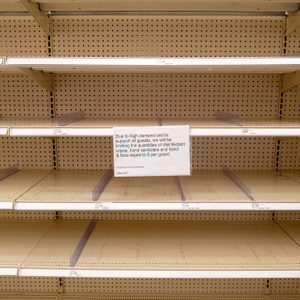 Empty shelves display a sign limiting disinfectant supply to six per guest, set in a store context with beige shelving.