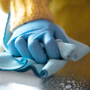 Gloved hand wipes glass surface with blue cloth, creating circular motion on soapy water, set against a blurred yellow background.
