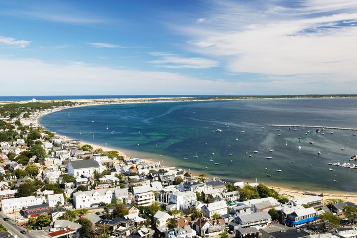 Boats float in a large bay beside a town with many houses. The scene includes a sandy shoreline and expansive blue sky.