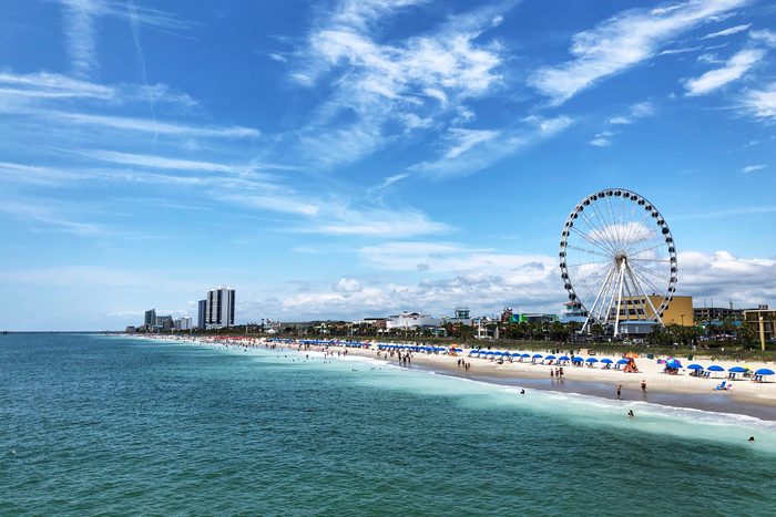 Ferris wheel stands tall, overlooking a sandy beach with umbrellas and people enjoying the sun, beside a cityscape of modern buildings under blue skies.