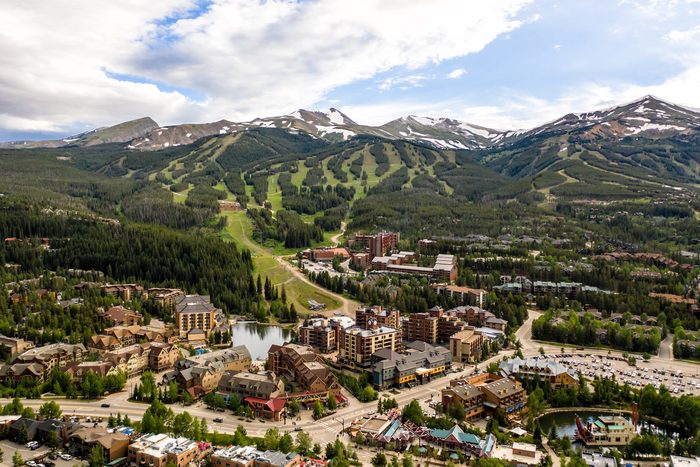 Buildings sit nestled among lush green trees, with ski slopes on mountainous terrain under a partly cloudy, blue sky backdrop.