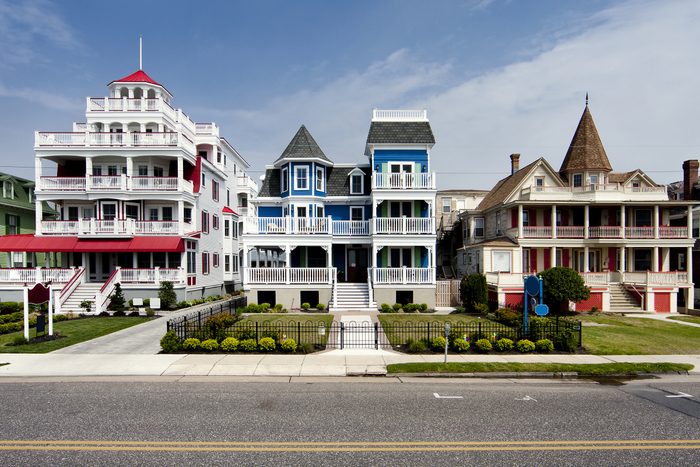 Three Victorian-style houses stand in a row, featuring colorful facades and ornate balconies, set against a clear sky and a well-kept front lawn.