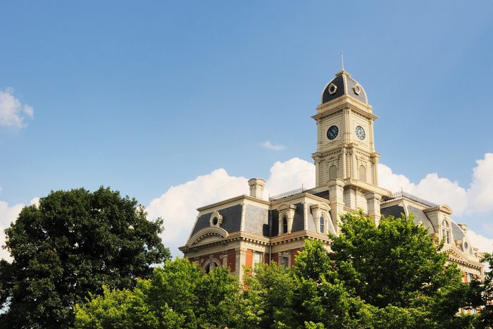 A clock tower rests atop a historic building surrounded by lush green trees under a bright blue sky.
