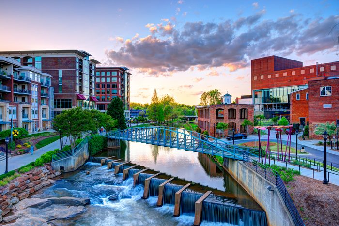 Bridge spans river, water cascades over small dam, surrounded by modern and historic brick buildings, trees, and a colorful sunset sky.