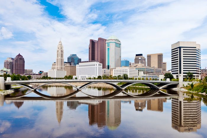 Skyline reflects on river beneath modern bridge, with diverse skyscrapers towering in background, under a bright, cloudy sky.