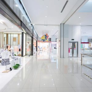 Mannequins display clothing in a brightly lit shopping mall corridor, surrounded by glossy floors and colorful decorations hanging from the ceiling.