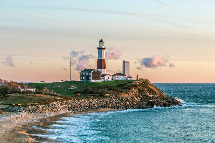 Lighthouse stands on a grassy hill above rocky shore, ocean waves beneath, with soft clouds and a pastel sky in the background.