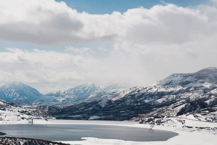 Snowy mountains stand quietly, surrounding a calm, partially frozen lake under a cloudy sky.