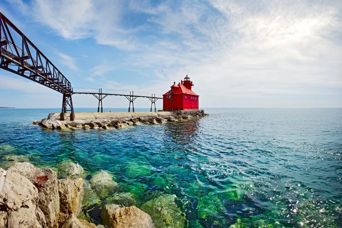 Red lighthouse stands on rocky pier, with a metal walkway extending towards it, surrounded by clear blue-green ocean under a cloudy sky.