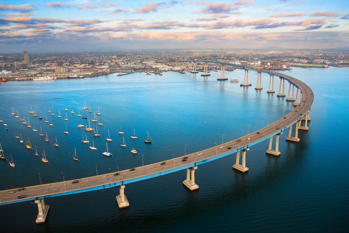 Bridge curves over blue water, with cars driving, surrounded by boats in a bustling city harbor under a partially cloudy sky.