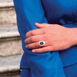 A hand displays a sapphire ring adorned with diamonds, placed gently on the other arm, wearing a blue sleeve, against stone steps.