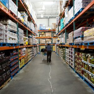 A person pushes a shopping cart down an aisle in a large warehouse, surrounded by shelves stocked with various boxed and palletized goods.