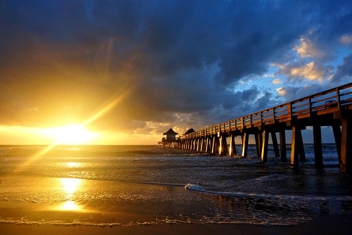 Sunset illuminates a long pier with dramatic clouds over a calm ocean, casting golden reflections on the wet sand.