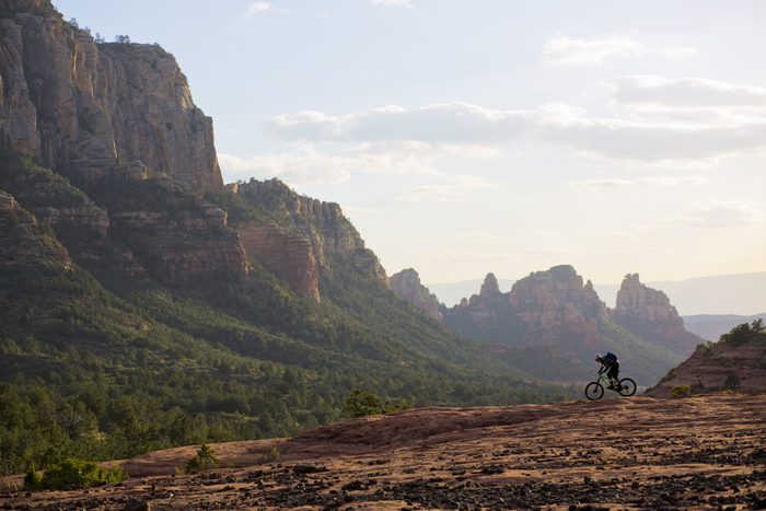 Bicyclist pedaling on rocky terrain, surrounded by a vast landscape of green valleys and towering cliffs under a partly cloudy sky.