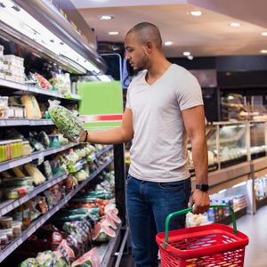 Man holds lettuce, examining it while shopping, in a grocery store with produce on shelves and a red basket in his other hand.