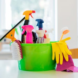 Cleaning supplies sit in a bright green bucket on a white counter, including spray bottles, brushes, yellow gloves, and a pink cloth.