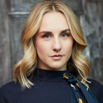A woman with blonde hair looks directly forward, wearing a dark top with a patterned scarf, standing against a dark, textured background.