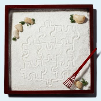 A red-framed sand tray holds rocks and a rake, featuring puzzle-piece patterns amidst scattered greenery on a light blue background.