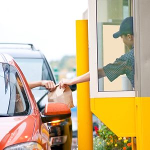 Customer receives paper bag through car window at drive-thru, served by an attendant behind a yellow booth.