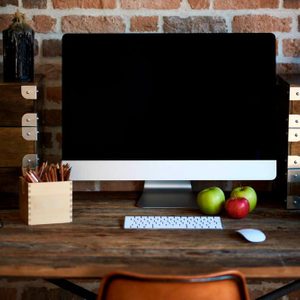 Computer monitor sits on a wooden desk, with keyboard, mouse, pencils, and apples nearby; surrounded by drawers and brick wall backdrop.