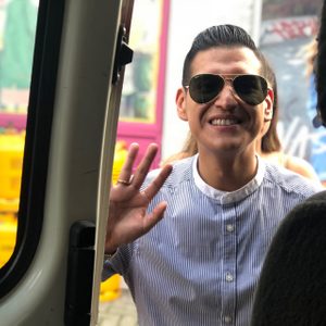 Person waving, wearing sunglasses and striped shirt, standing outside a partially open door; bright urban setting with graffiti and pink-framed window.