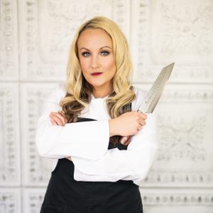 A person in a chef's uniform holds a knife, posed confidently against an ornate, patterned white wall.
