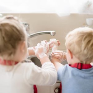 Children washing hands with soap, standing at a kitchen sink, sunlight filtering through a window.