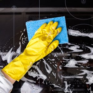 A gloved hand cleans a stovetop with a blue sponge, surrounded by soap suds.