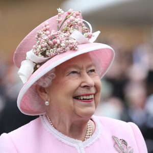 A person in a pink floral hat and matching coat smiles warmly, with a blurred crowd in the background.