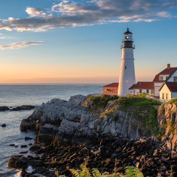 A lighthouse stands atop rocky cliffs, overlooking the ocean at sunset, with a house nearby and a clear sky above.