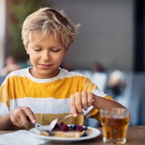Child eating pancakes with berries, indoors at a restaurant, near a glass of amber juice.
