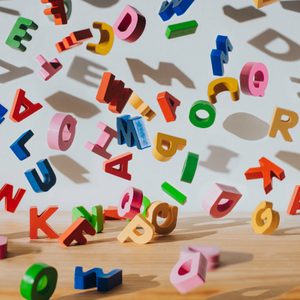 Colorful wooden letters scatter across a light wooden surface, casting shadows on a plain background.