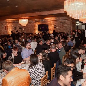 Guests dine and socialize at long tables in a warmly lit restaurant with decorative brick walls and chandeliers.