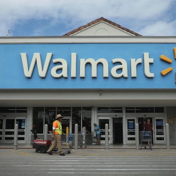 Storefront displays "Walmart" sign; people push carts outside, one worker passes with cart. Overcast sky above.