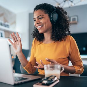 A woman wearing headphones smiles and waves at a laptop, sitting at a table with a glass mug and smartphone in a cozy room.