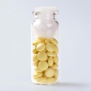 A glass bottle holds yellow, round tablets with white cotton on top, set against a plain, light gray background.