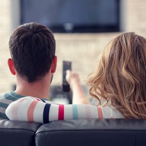 Two people sit on a couch, watching television, with a remote control in hand, in a cozy living room setting.
